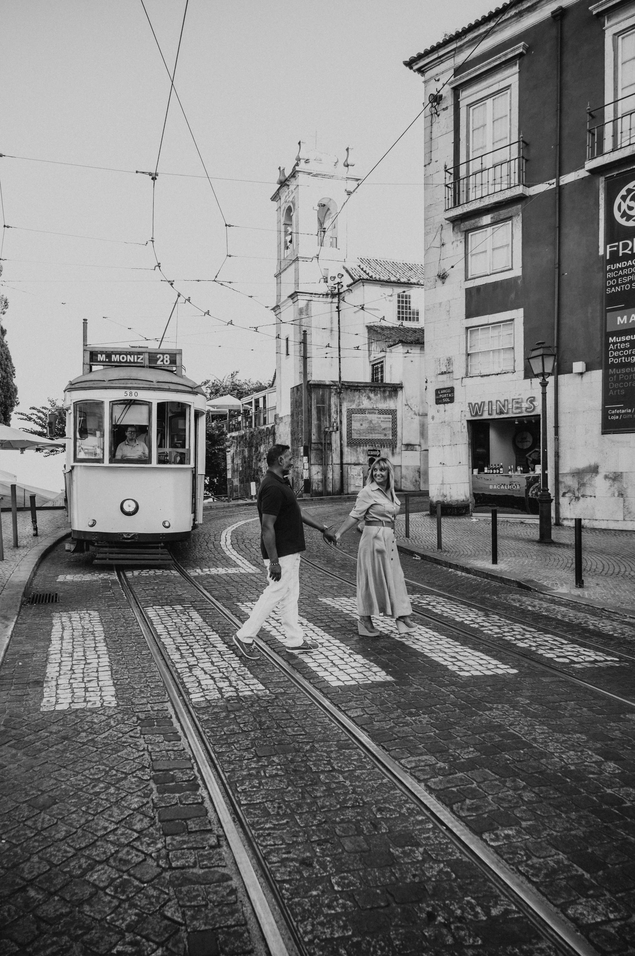 Black-and-white photograph: the couple standing together beside a Lisbon street with a yellow tram behind them during their couple photoshoot in Lisbon.