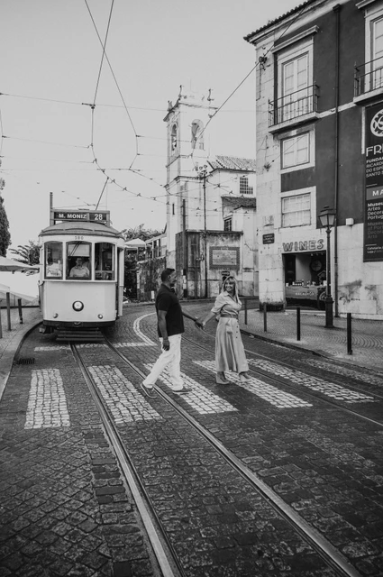 Black-and-white photograph: the couple standing together beside a Lisbon street with a yellow tram behind them during their couple photoshoot in Lisbon.