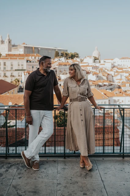 A shared smile above Lisbon’s rooftops | Lisbon couple photoshoot The couple smiling at each other beside the viewpoint railing with Lisbon’s rooftops behind them during their couple photoshoot in Lisbon.