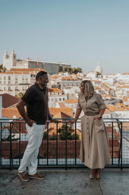 A shared smile above Lisbon’s rooftops | Lisbon couple photoshoot The couple smiling at each other beside the viewpoint railing with Lisbon’s rooftops behind them during their couple photoshoot in Lisbon.