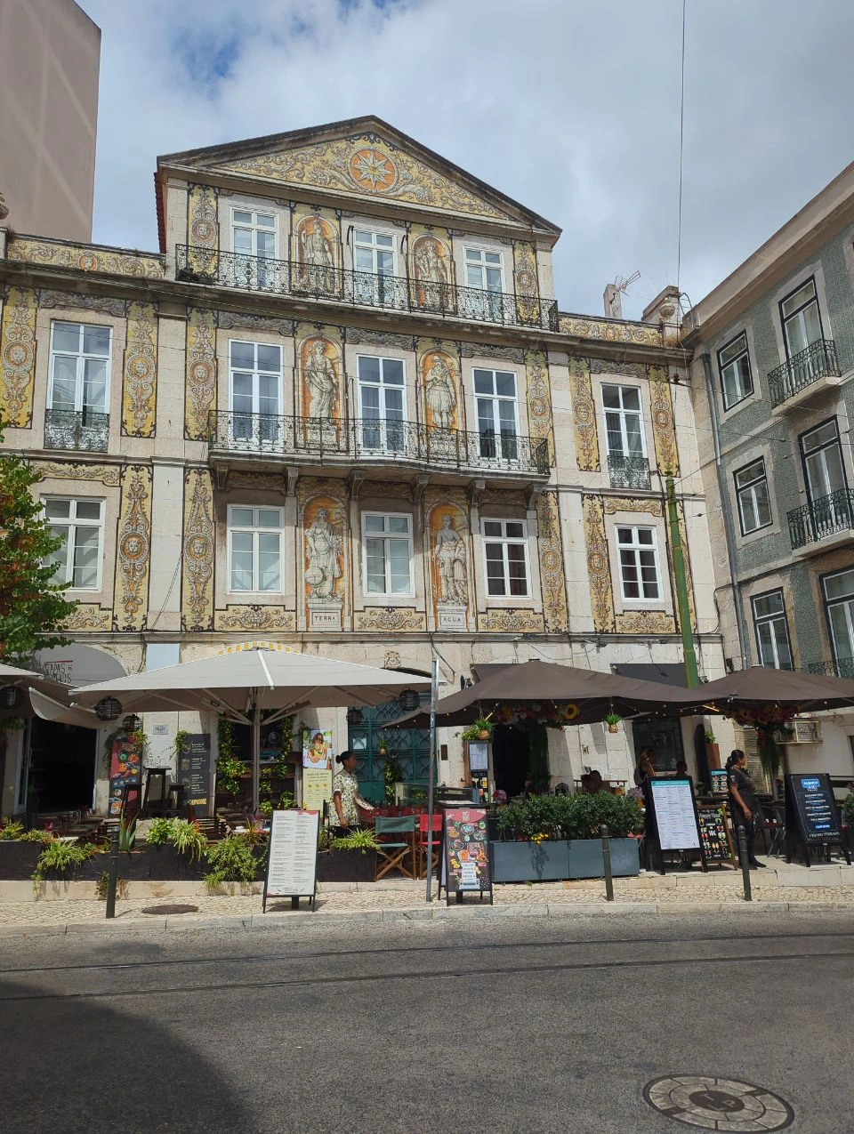 Lisbon Photoshoot Route - Ornate Tiled Facade An ornate Lisbon facade covered in figurative ceramic tiles rises above restaurant awnings beside a street with rails.