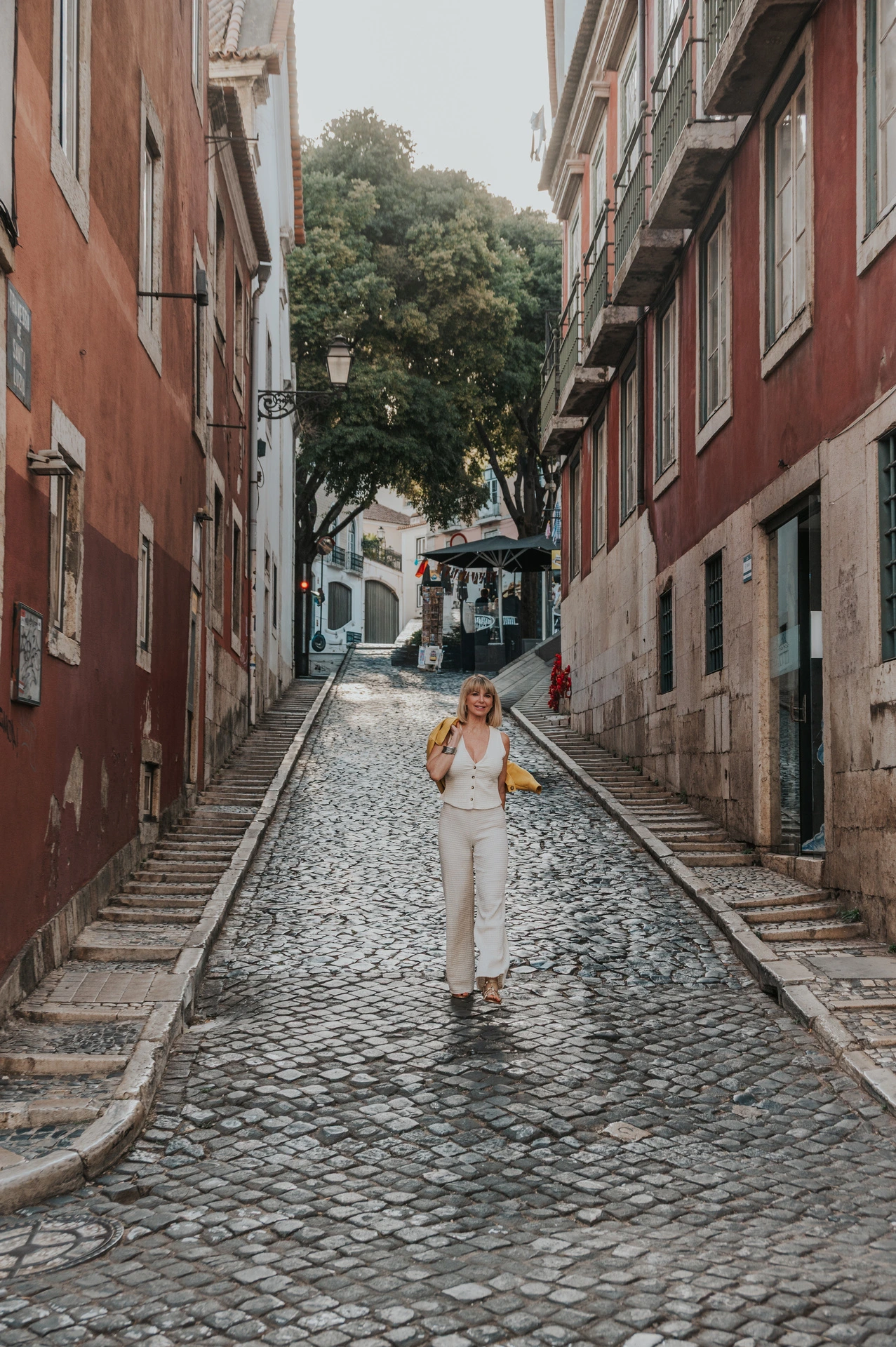 A distant view of the woman walking down a cobbled street between red buildings in Alfama during her solo photoshoot in Lisbon.