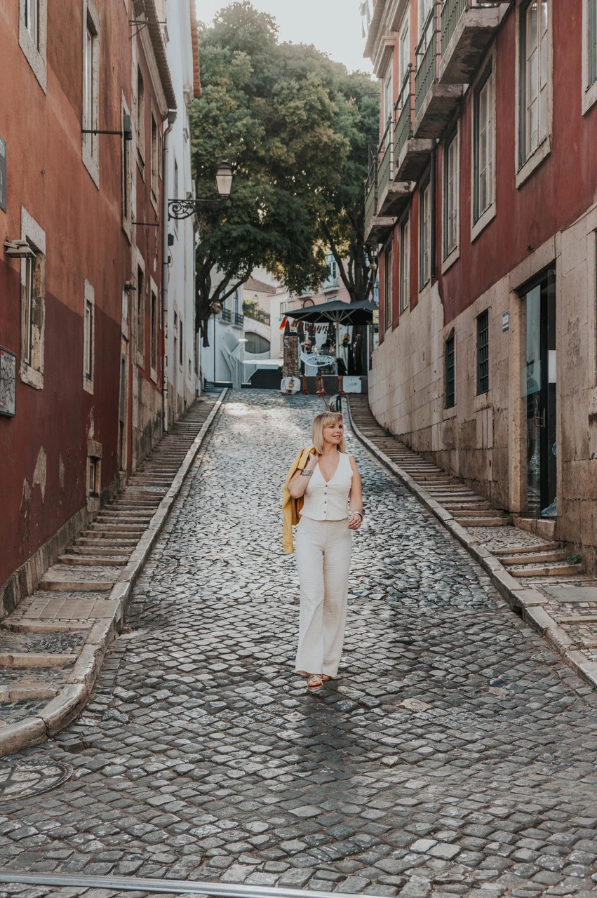 A street portrait in Alfama | Lisbon solo photoshoot The woman walking towards the camera along a cobbled Alfama street with her yellow jacket over one shoulder during her solo photoshoot in Lisbon.