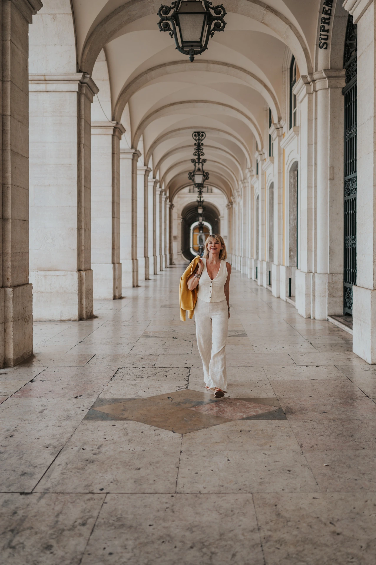 The woman walking beneath the repeated arches and hanging lanterns at Praça do Comércio during her solo photoshoot in Lisbon.
