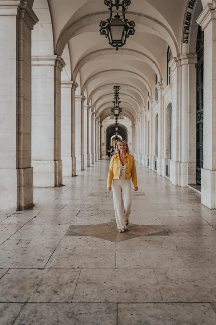 A yellow jacket beneath the arcade | Lisbon solo photoshoot The woman holding her yellow jacket over one shoulder as she walks beneath the arcade at Praça do Comércio during her solo photoshoot in Lisbon.