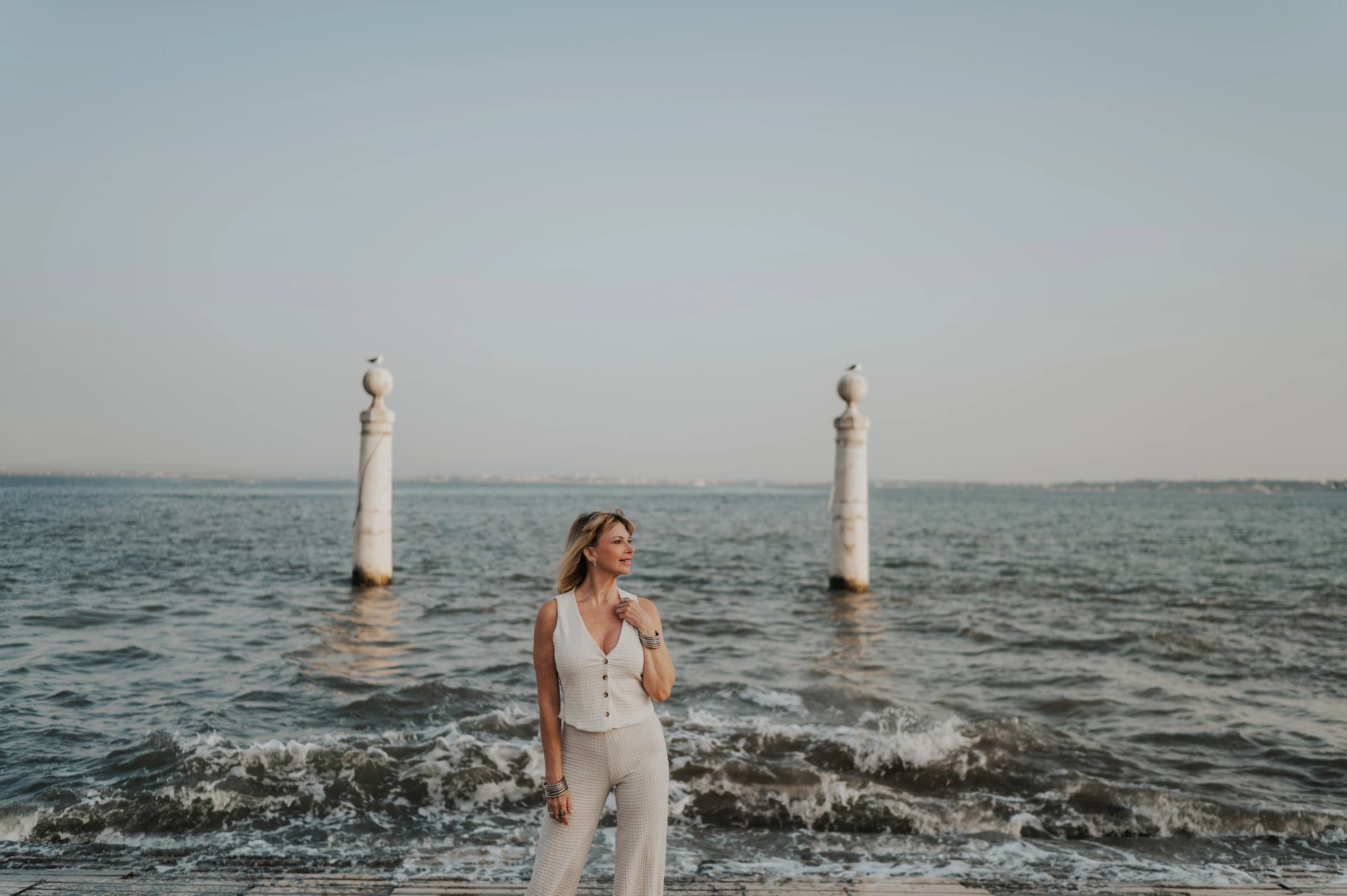 A wide portrait of the woman by the water with waves breaking behind her near Praça do Comércio during her solo photoshoot in Lisbon.