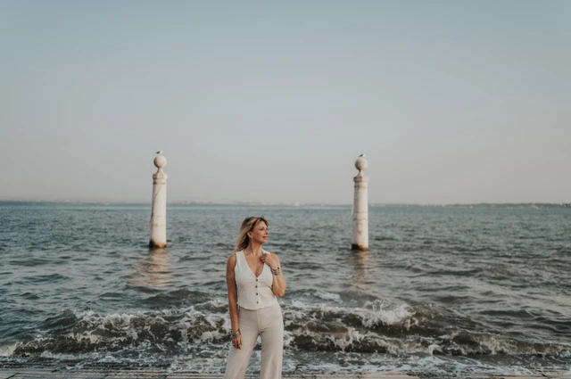 A wide portrait of the woman by the water with waves breaking behind her near Praça do Comércio during her solo photoshoot in Lisbon.