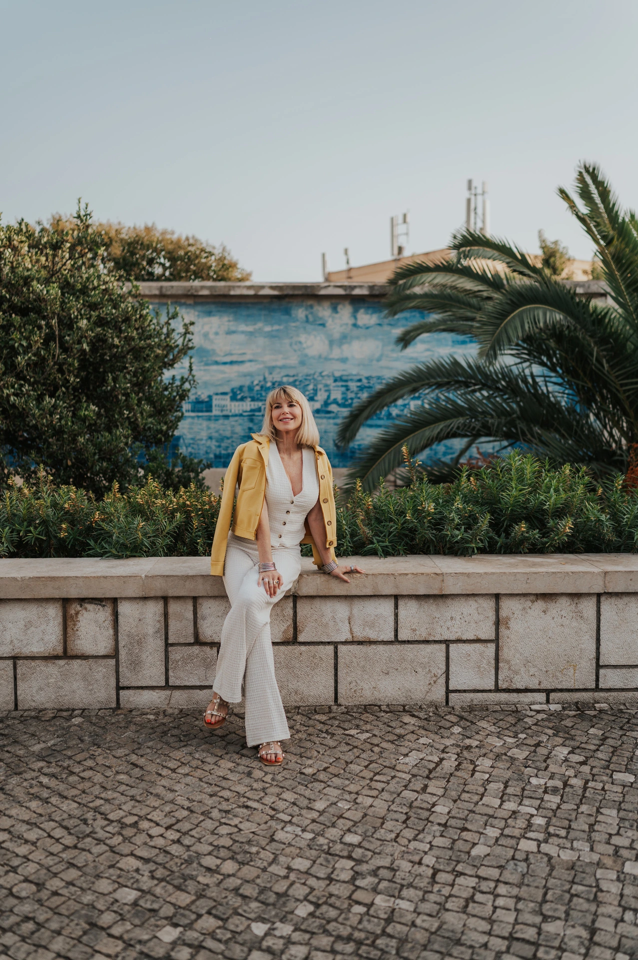 The woman smiling while seated on the low garden wall beneath the palms at Santa Luzia during her solo photoshoot in Lisbon.
