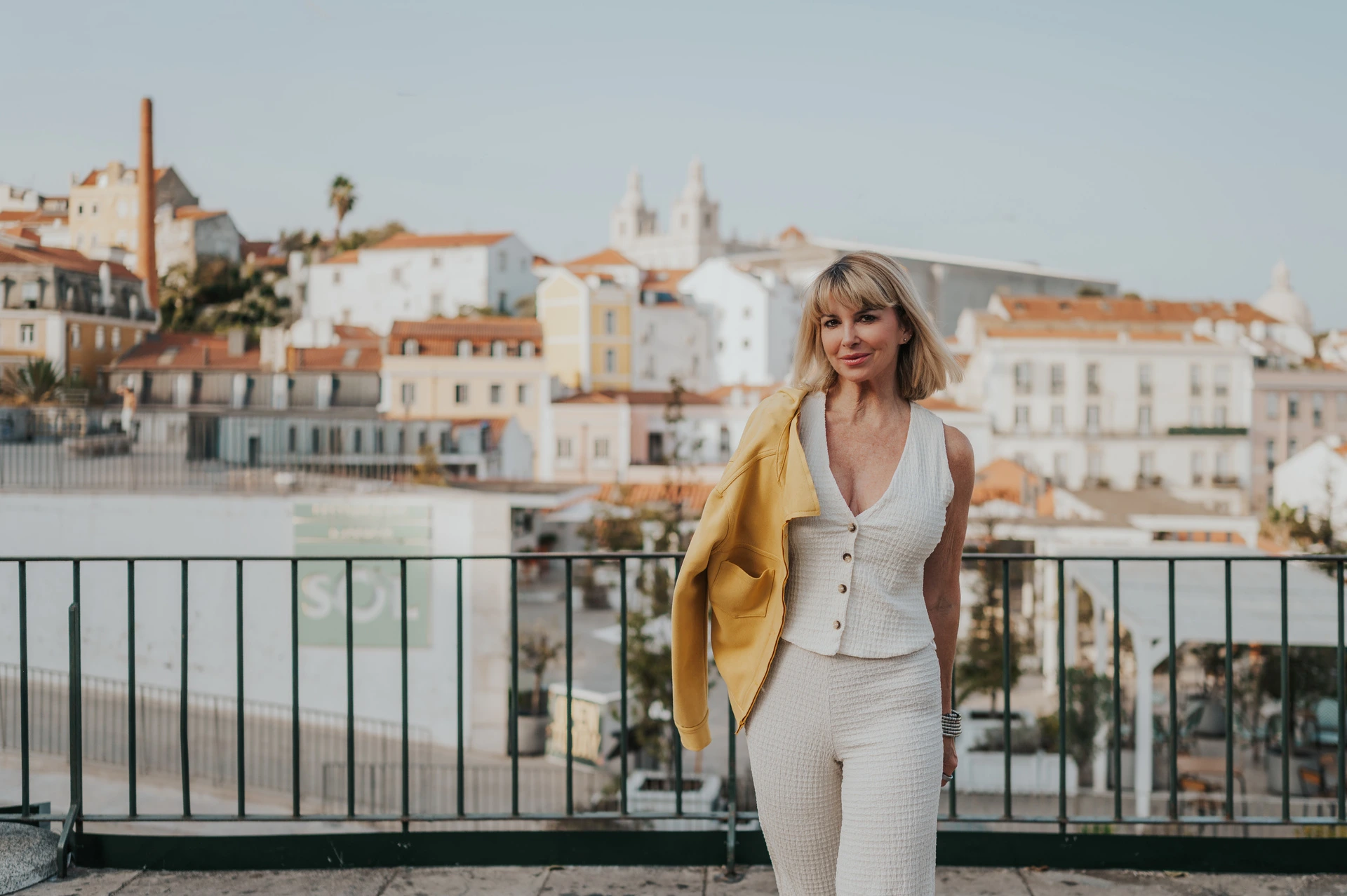 A wide rooftop portrait at Portas do Sol | Lisbon solo photoshoot A wide portrait of the woman with a yellow jacket over her shoulder and Alfama’s rooftops behind her during her solo photoshoot in Lisbon.