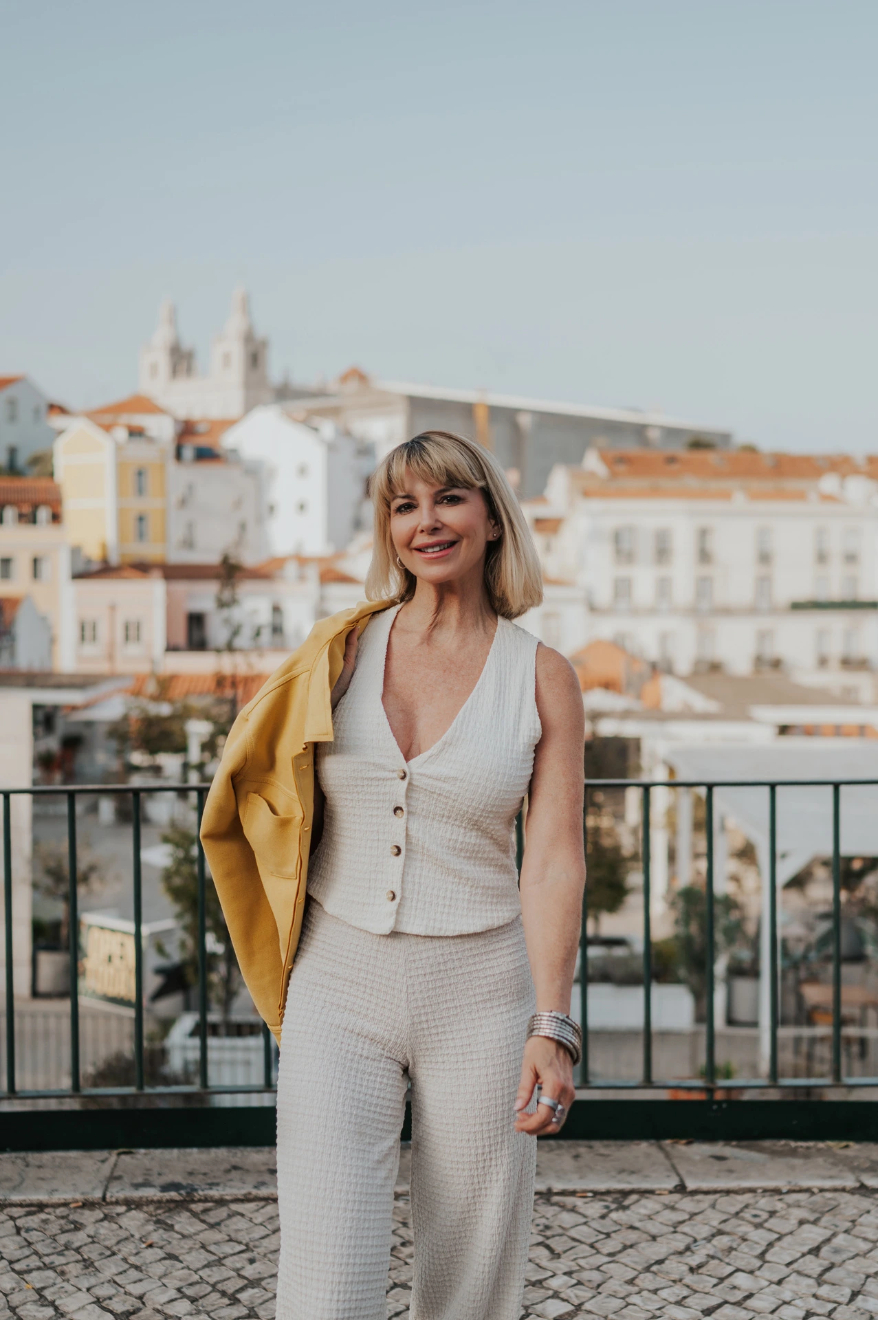 The woman smiling with a yellow jacket over one shoulder above the rooftops at Portas do Sol during her solo photoshoot in Lisbon.
