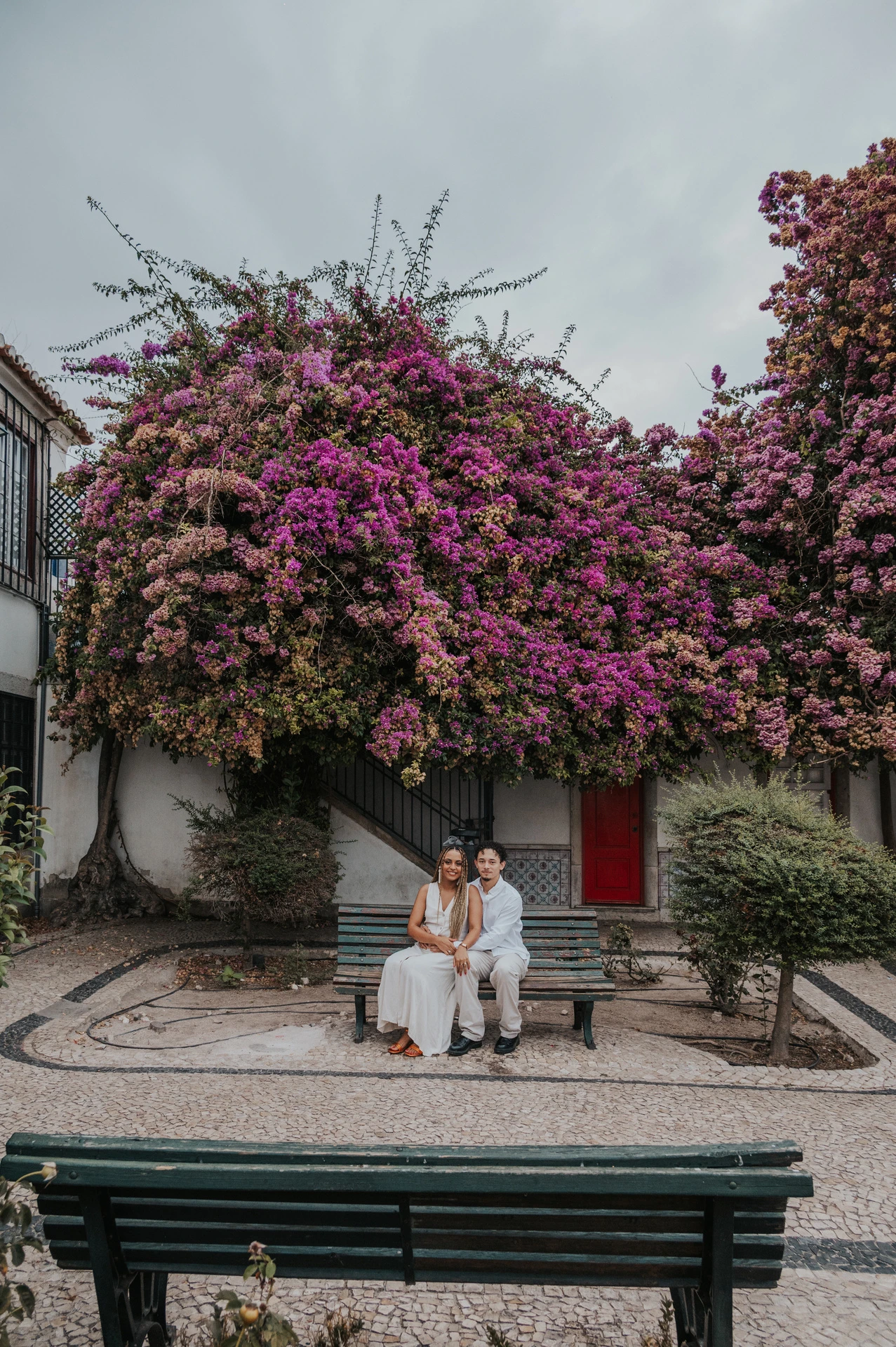 The couple seated together on a garden bench beneath flowering branches during their couple photoshoot in Lisbon.