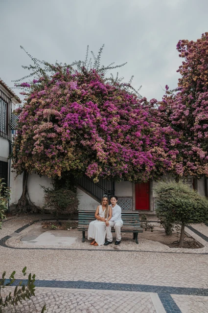 A seated portrait beneath the flowers | Lisbon couple photoshoot The couple seated together on a garden bench beneath flowering branches during their couple photoshoot in Lisbon.