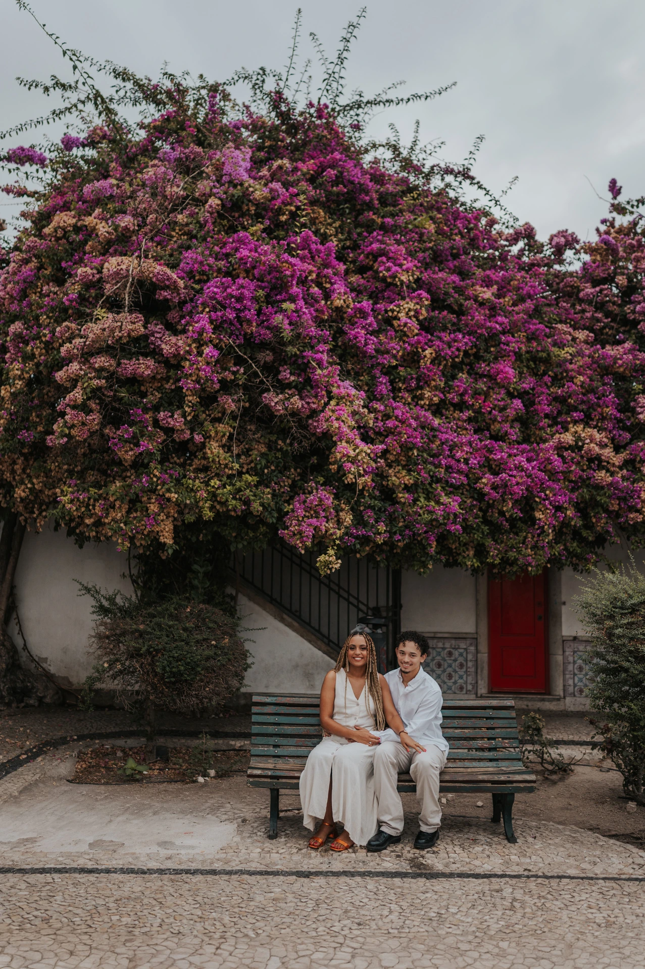 The couple seated together on a garden bench beneath flowering branches during their couple photoshoot in Lisbon.