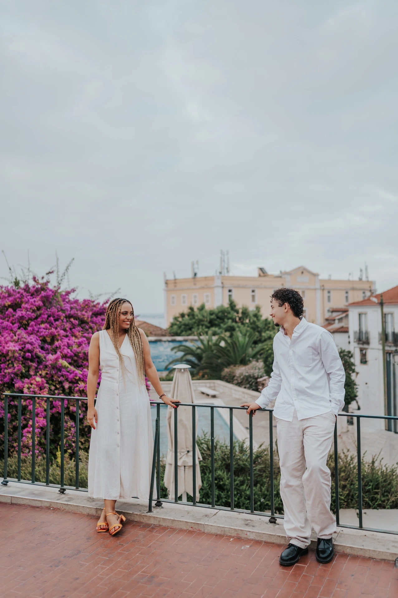 The couple smiling at each other beside the garden railing and purple flowers during their couple photoshoot in Lisbon.