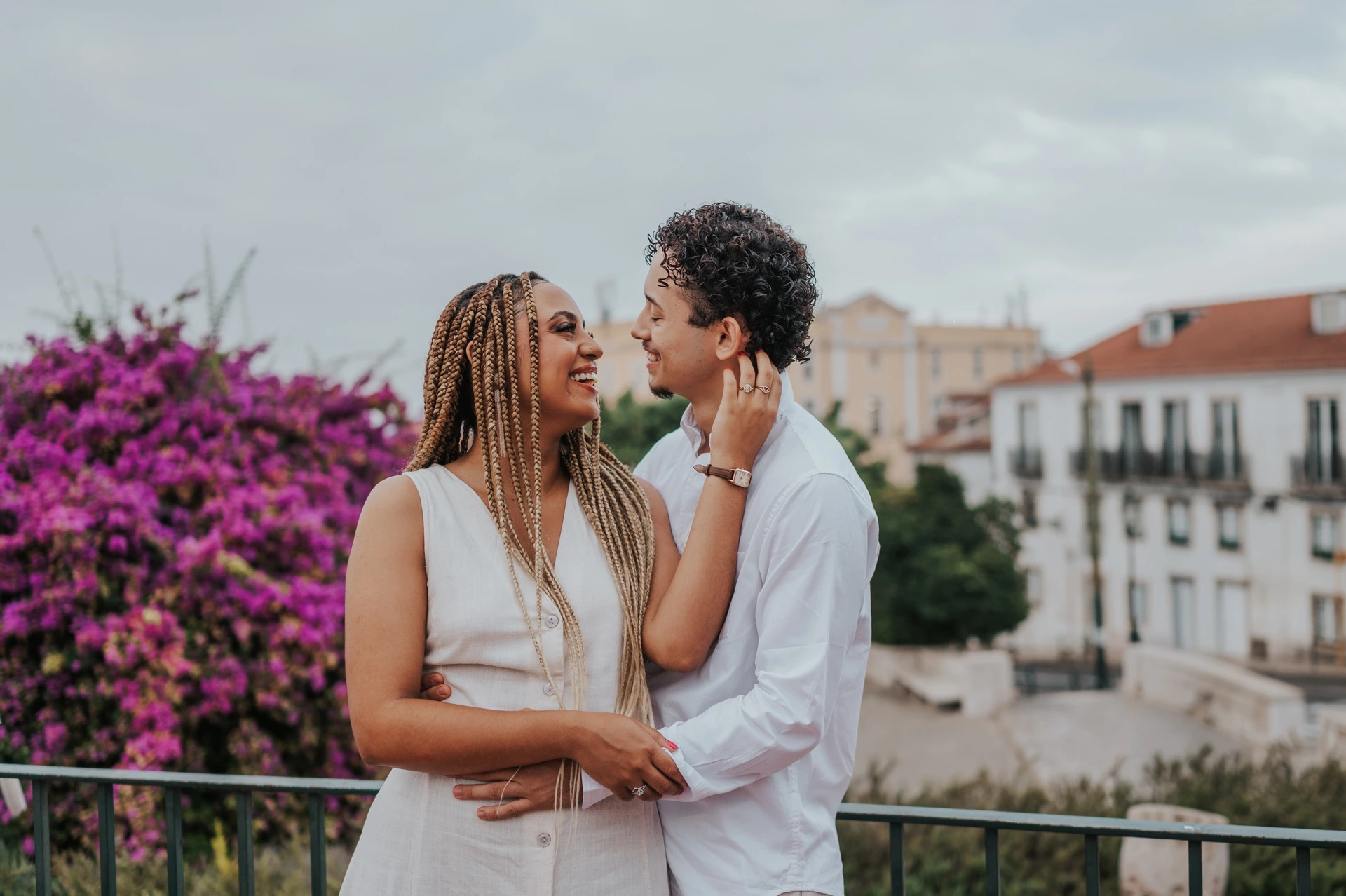 The couple smiling at each other beside the garden railing and purple flowers during their couple photoshoot in Lisbon.