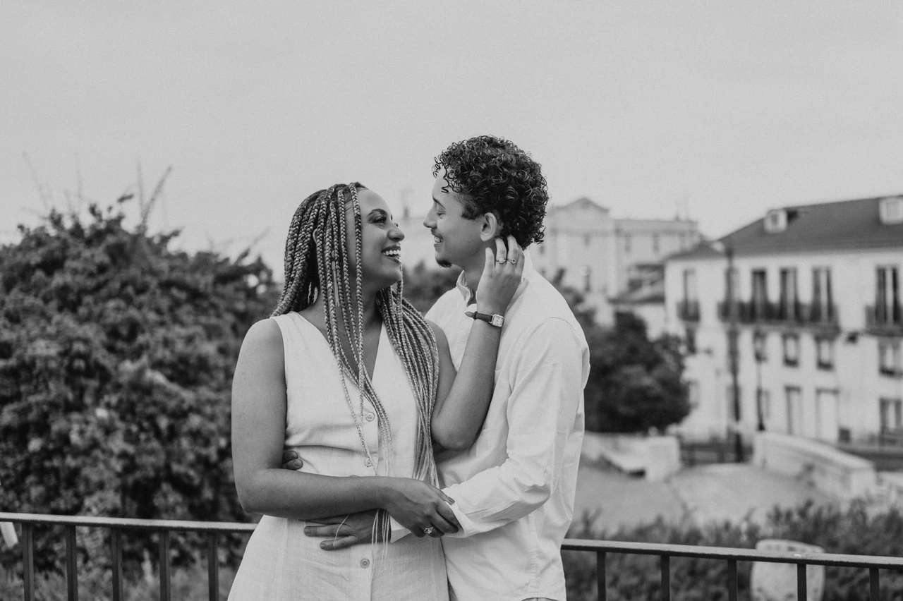 A shared smile beside the flowers in black and white | Lisbon couple photoshoot Black-and-white photograph: the couple smiling at each other beside the garden railing and purple flowers during their couple photoshoot in Lisbon.