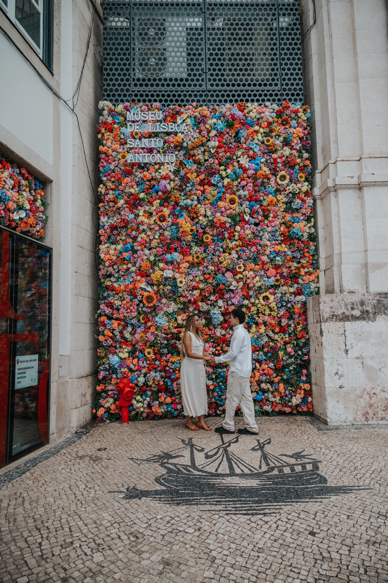The couple standing close together in front of a wall covered in colourful flowers during their couple photoshoot in Lisbon.