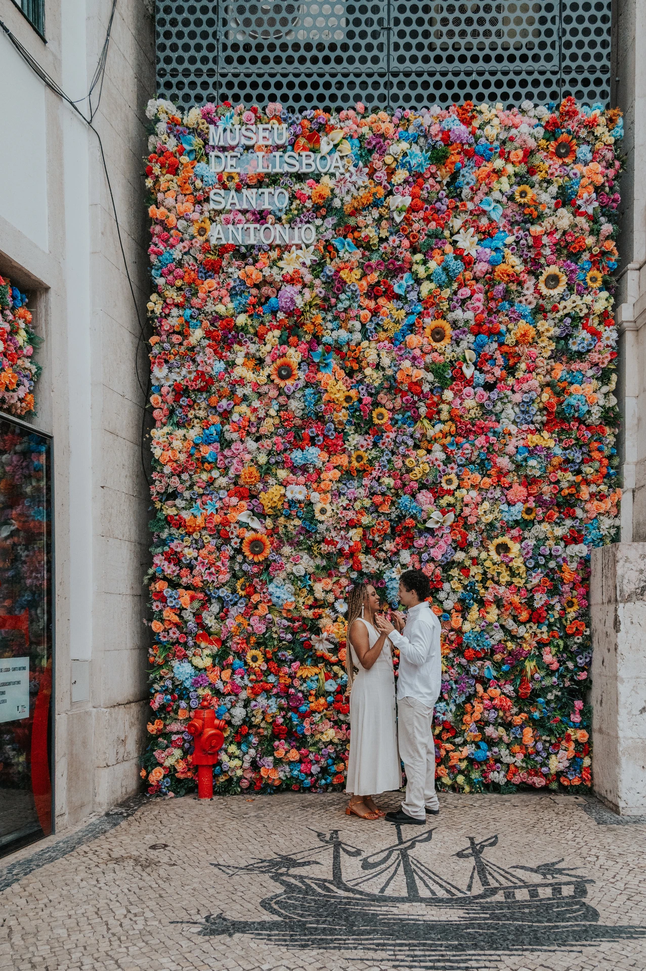 The couple standing close together in front of a wall covered in colourful flowers during their couple photoshoot in Lisbon.