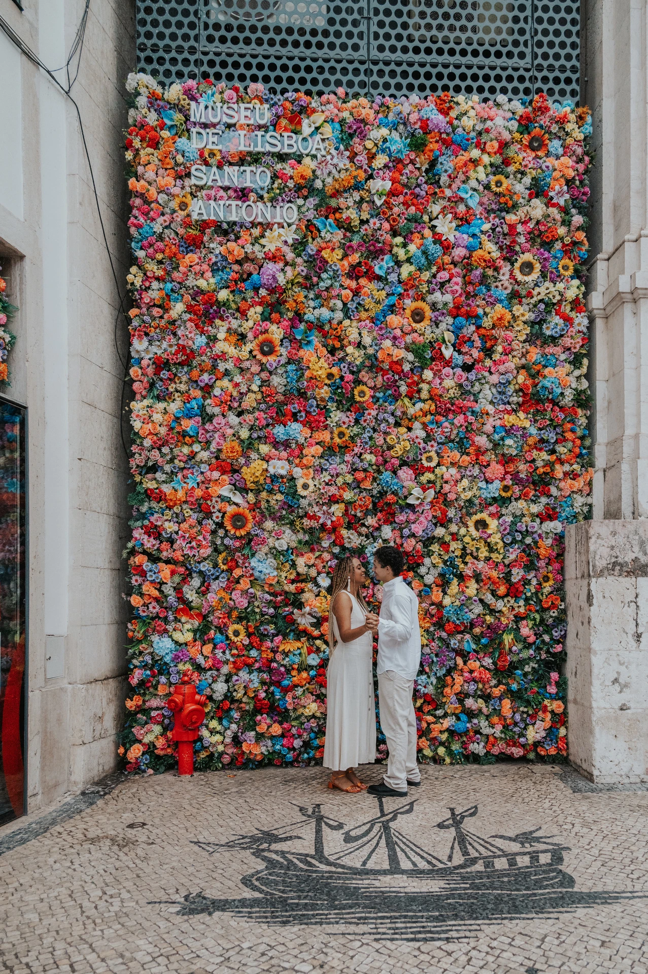 The couple standing close together in front of a wall covered in colourful flowers during their couple photoshoot in Lisbon.
