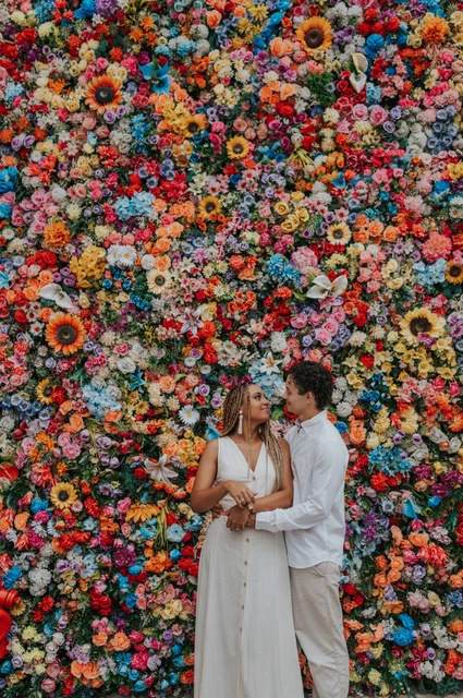 Together against the colourful flower wall | Lisbon couple photoshoot The couple standing close together in front of a wall covered in colourful flowers during their couple photoshoot in Lisbon.