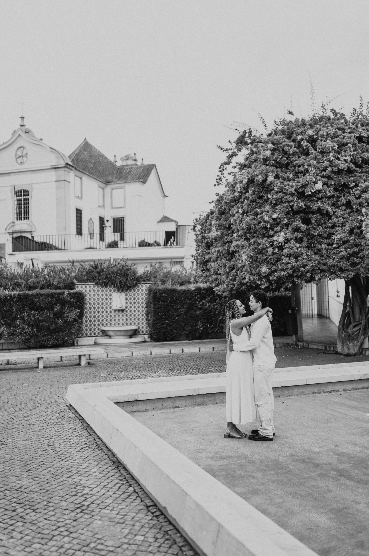 Black-and-white photograph: the couple embracing beside the shallow garden pool and flowering branches during their couple photoshoot in Lisbon.
