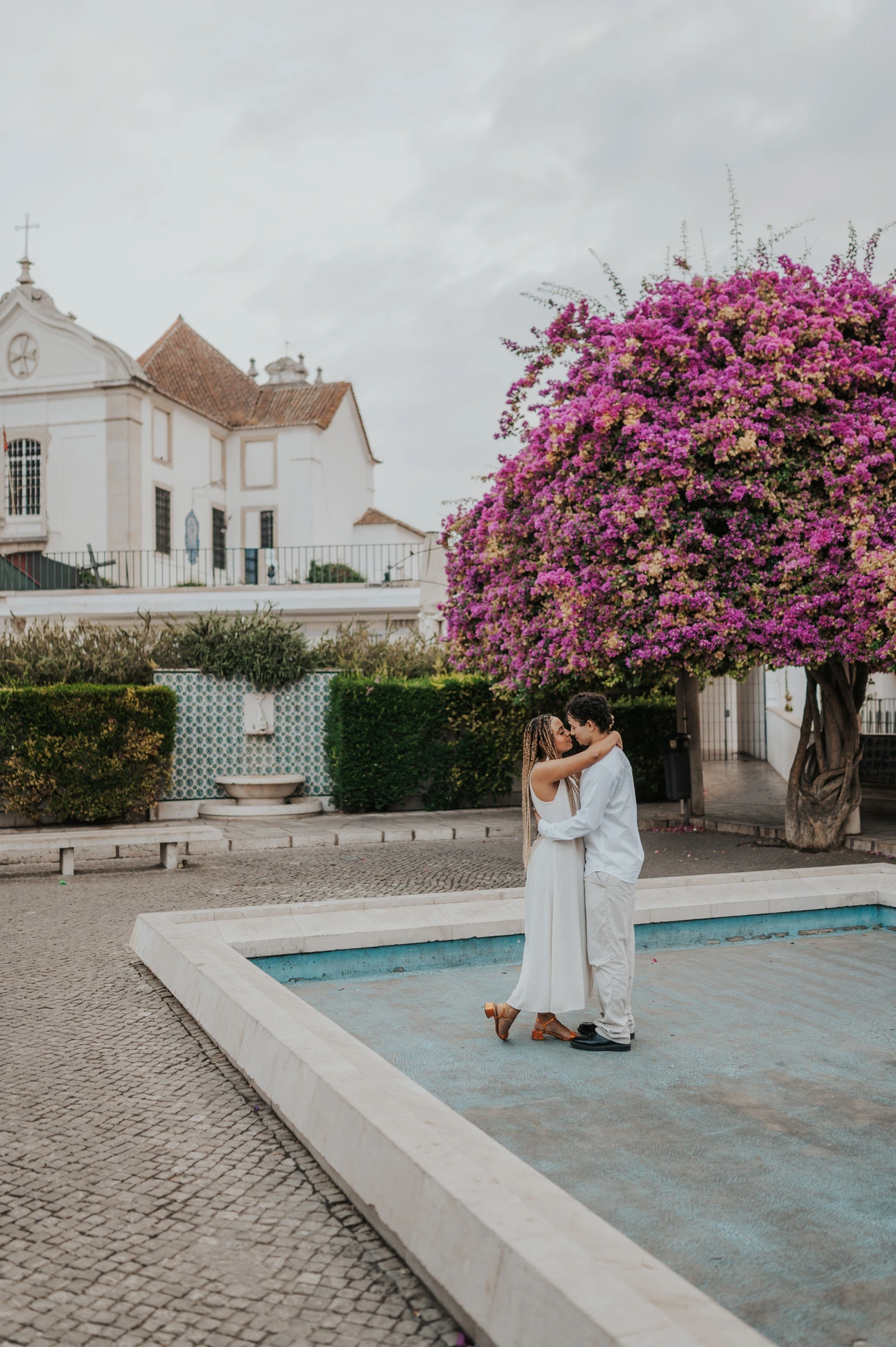 The couple embracing beside the shallow garden pool and flowering branches during their couple photoshoot in Lisbon.