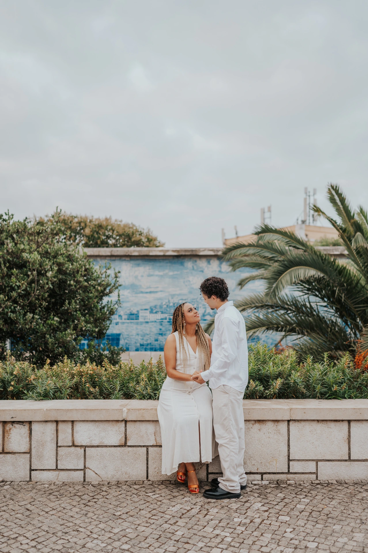 The woman seated on a low wall with her partner close beside her by the palm leaves during their couple photoshoot in Lisbon.