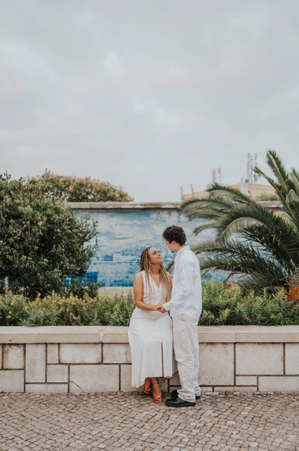 A close portrait beside the palm-lined viewpoint | Lisbon couple photoshoot The woman seated on a low wall with her partner close beside her by the palm leaves during their couple photoshoot in Lisbon.