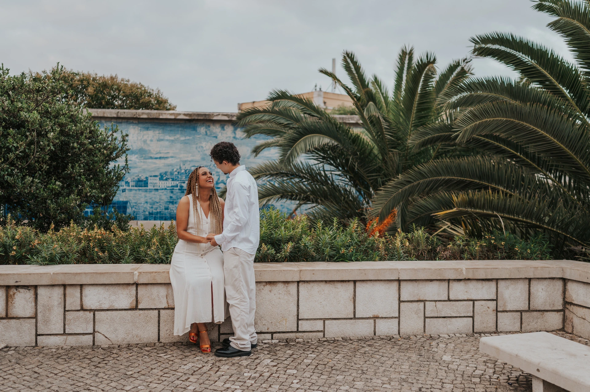 The woman seated on a low wall with her partner close beside her by the palm leaves during their couple photoshoot in Lisbon.