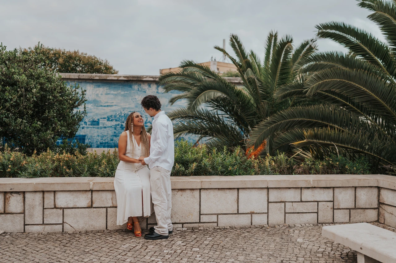 A close portrait beside the palm-lined viewpoint | Lisbon couple photoshoot The woman seated on a low wall with her partner close beside her by the palm leaves during their couple photoshoot in Lisbon.