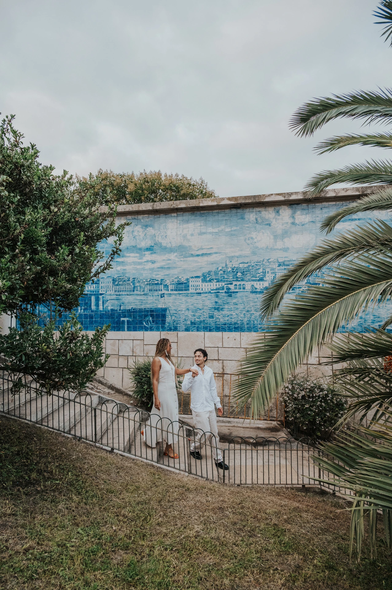The couple holding hands beside a blue tile panel and palm leaves at the viewpoint during their couple photoshoot in Lisbon.