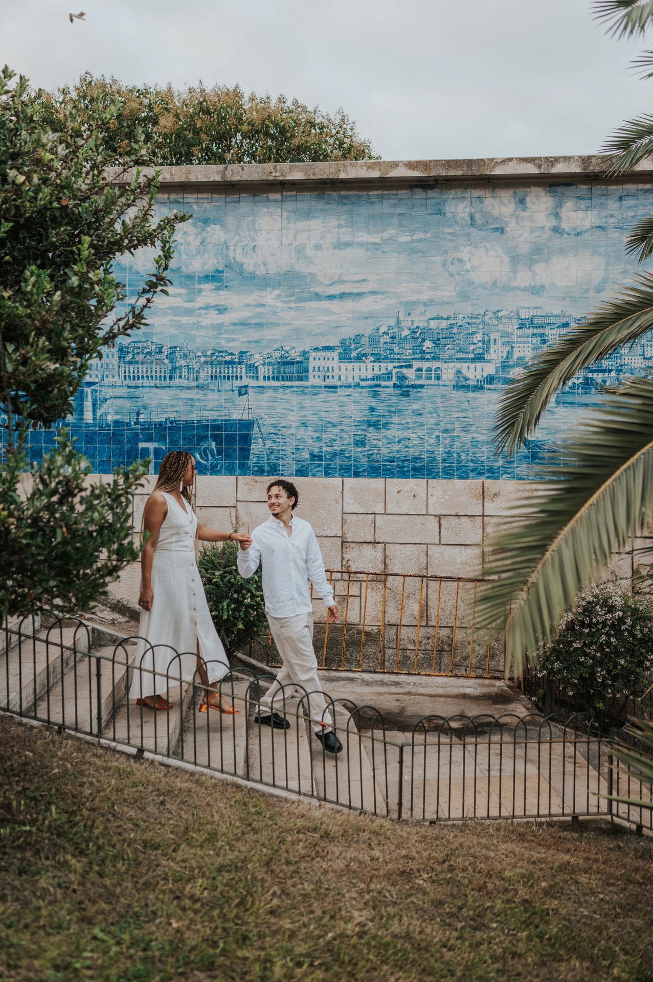 The couple holding hands beside a blue tile panel and palm leaves at the viewpoint during their couple photoshoot in Lisbon.