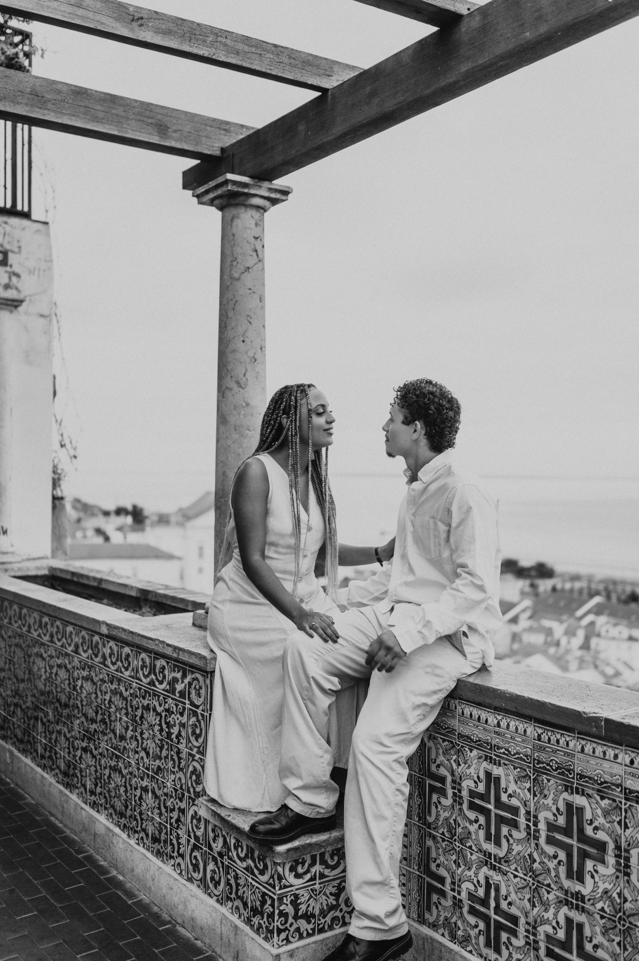 Black-and-white photograph: the couple seated facing each other on the tiled wall beneath the pergola during their couple photoshoot in Lisbon.