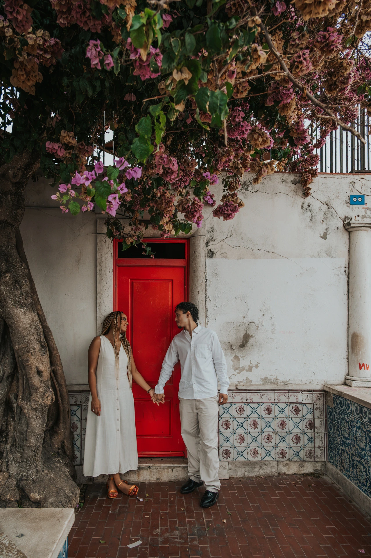 The couple holding hands on either side of a red door beneath flowering branches during their couple photoshoot in Lisbon.