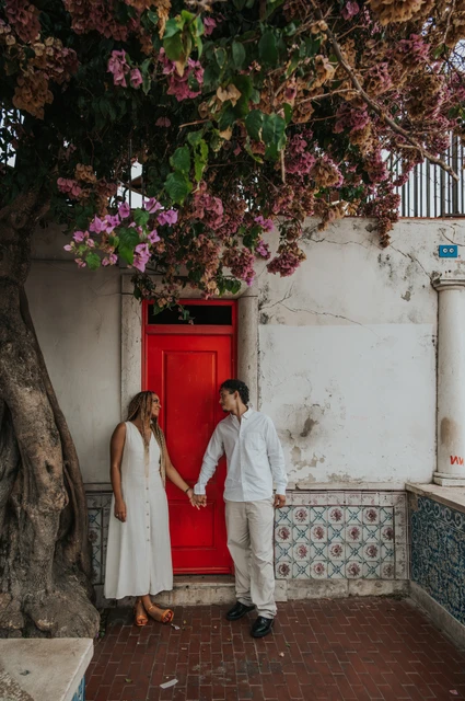 Joined hands beside the red door | Lisbon couple photoshoot The couple holding hands on either side of a red door beneath flowering branches during their couple photoshoot in Lisbon.