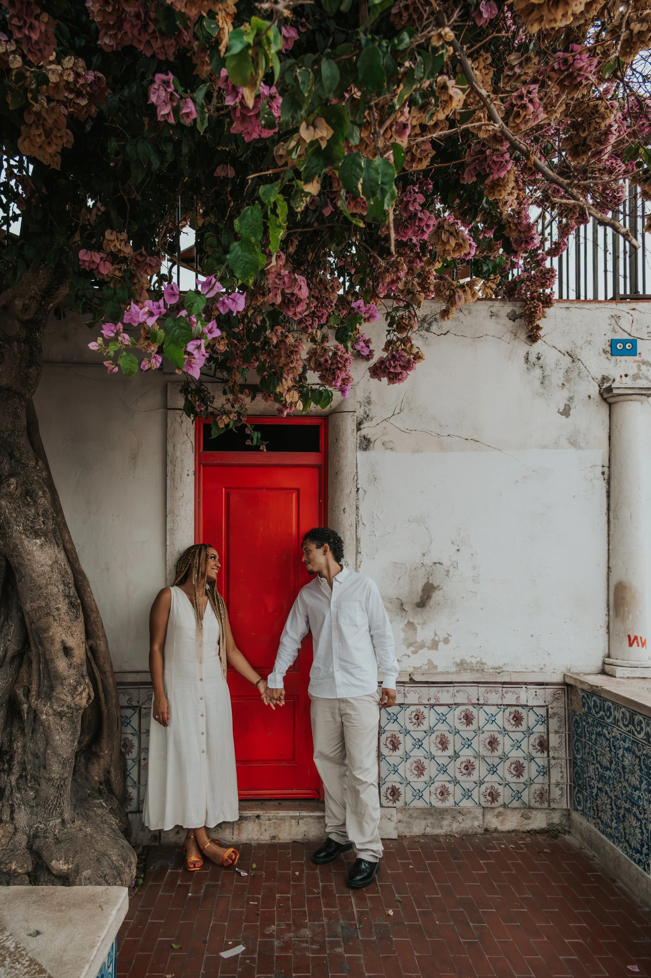 The couple holding hands on either side of a red door beneath flowering branches during their couple photoshoot in Lisbon.