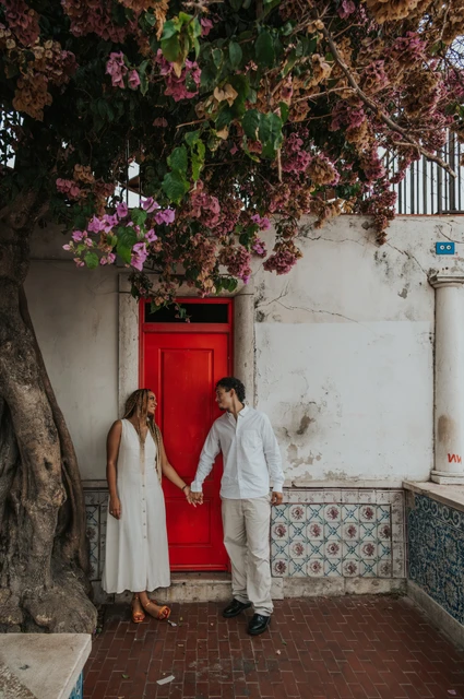 Joined hands beside the red door | Lisbon couple photoshoot The couple holding hands on either side of a red door beneath flowering branches during their couple photoshoot in Lisbon.