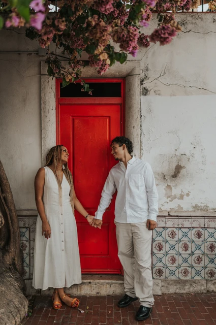 Joined hands beside the red door | Lisbon couple photoshoot The couple holding hands on either side of a red door beneath flowering branches during their couple photoshoot in Lisbon.