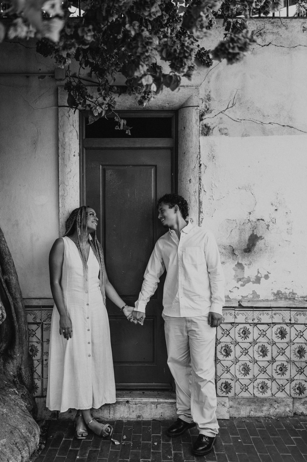 Black-and-white photograph: the couple holding hands on either side of a red door beneath flowering branches during their couple photoshoot in Lisbon.