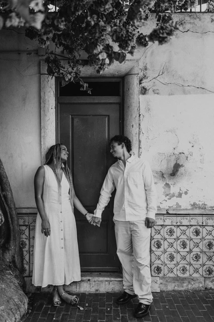 Joined hands beside the red door in black and white | Lisbon couple photoshoot Black-and-white photograph: the couple holding hands on either side of a red door beneath flowering branches during their couple photoshoot in Lisbon.