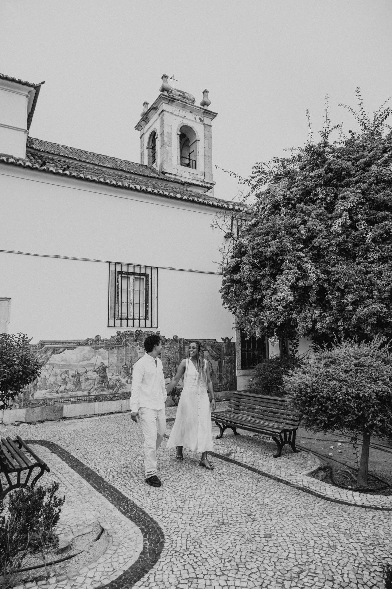 Black-and-white photograph: the couple holding hands along the garden path by Igreja de Santa Luzia and its blue tile panels during their couple photoshoot in Lisbon.