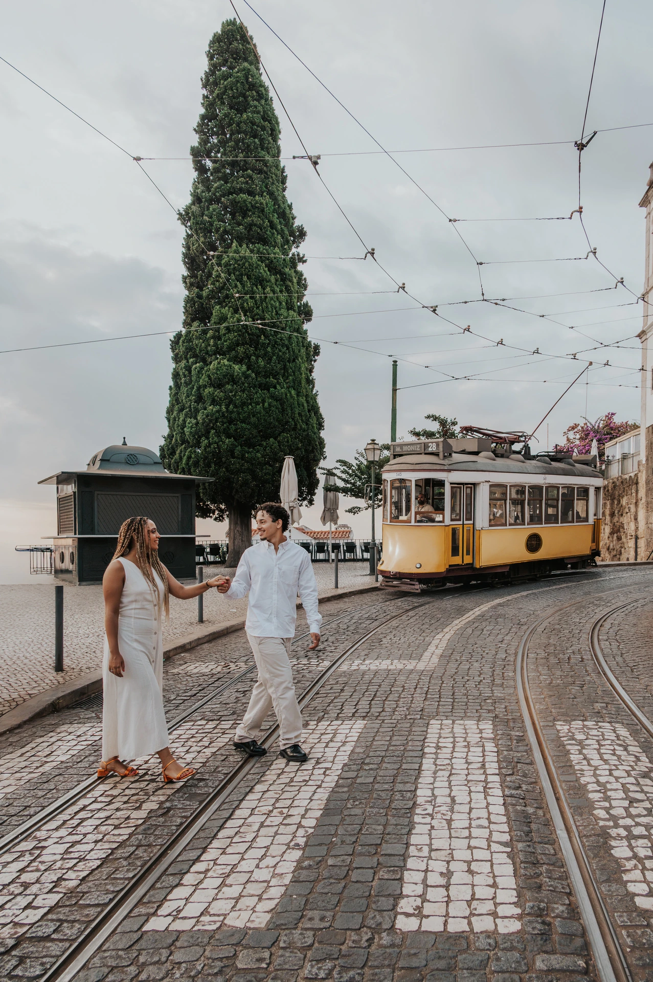 The couple walking together beside the curved tram tracks with a yellow tram in the background during their couple photoshoot in Lisbon.