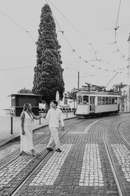 A street walk beside the yellow tram in black and white | Lisbon couple photoshoot Black-and-white photograph: the couple walking together beside the curved tram tracks with a yellow tram in the background during their couple photoshoot in Lisbon.