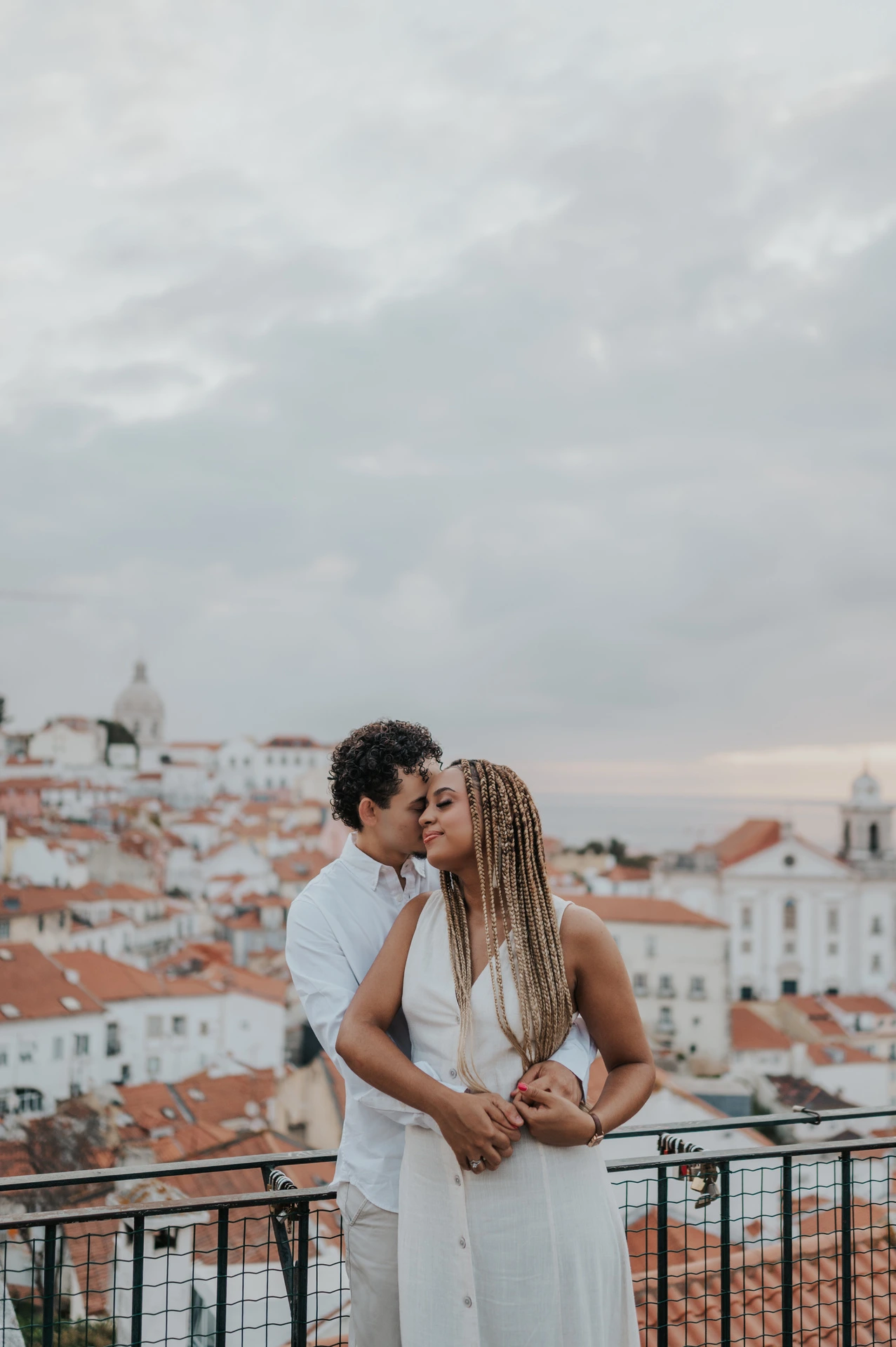 The man embracing his partner with the red rooftops and water behind them at the viewpoint during their couple photoshoot in Lisbon.