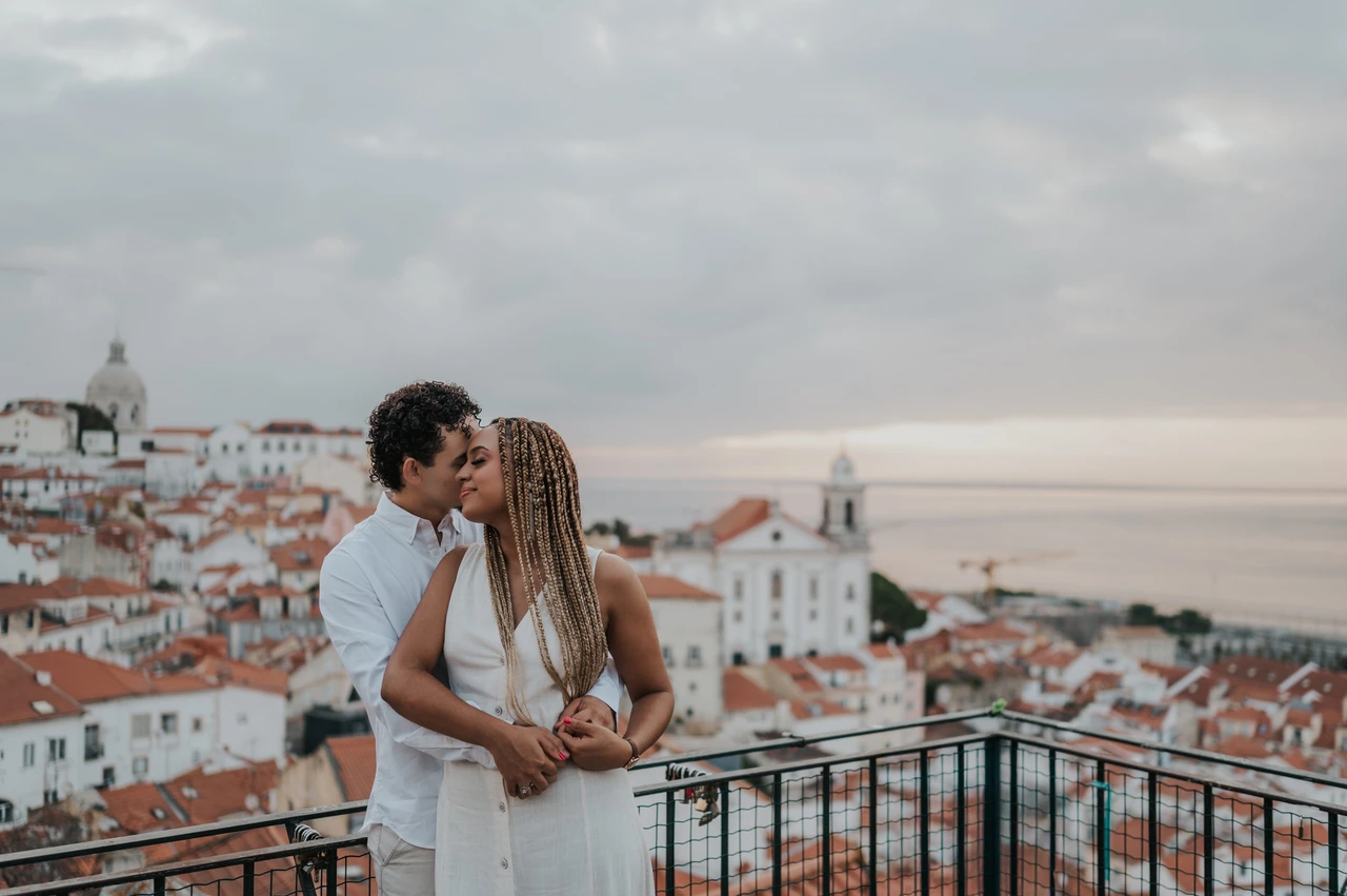 An embrace above the city | Lisbon couple photoshoot The man embracing his partner with the red rooftops and water behind them at the viewpoint during their couple photoshoot in Lisbon.