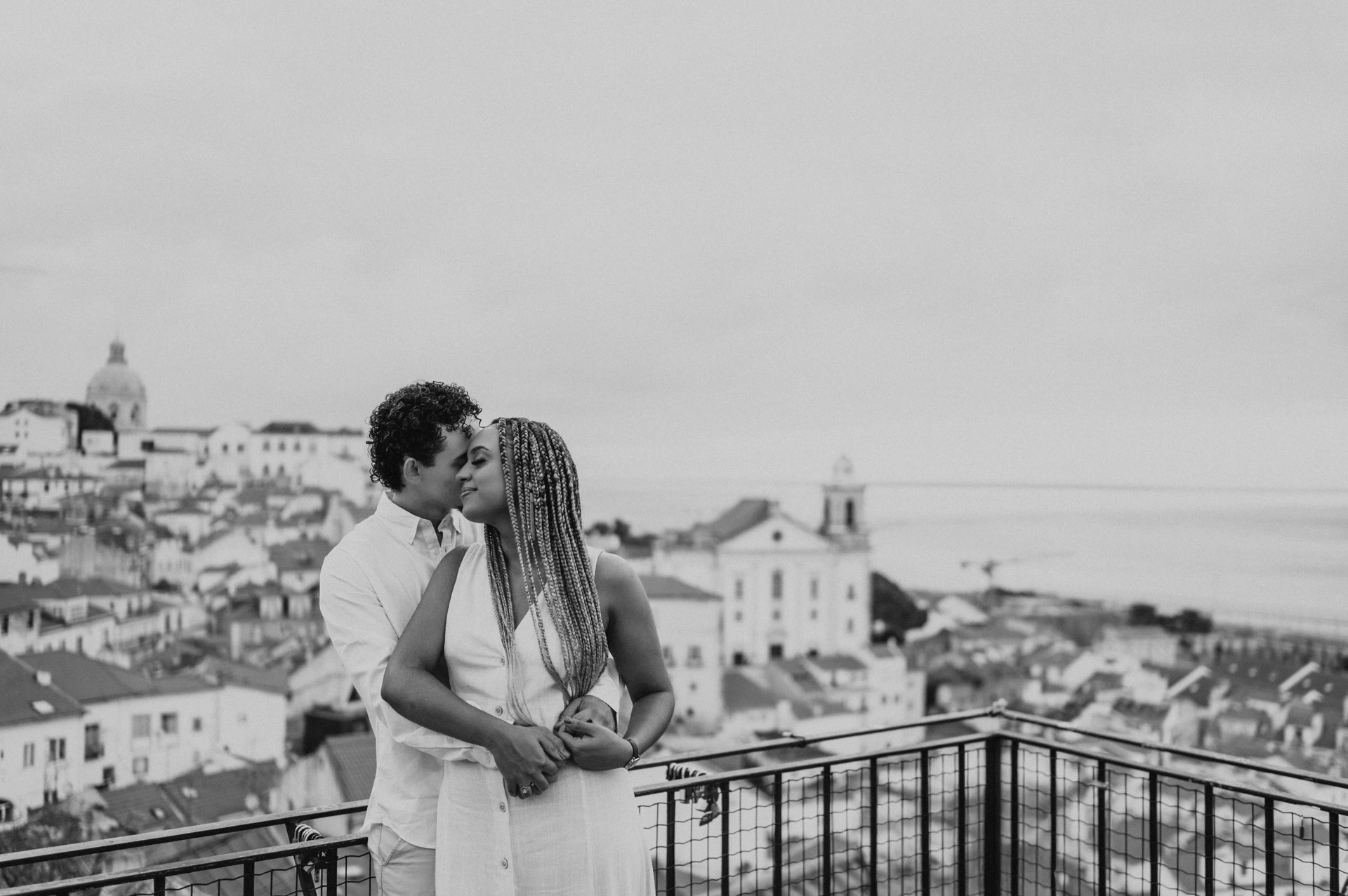Black-and-white photograph: the man embracing his partner with the red rooftops and water behind them at the viewpoint during their couple photoshoot in Lisbon.