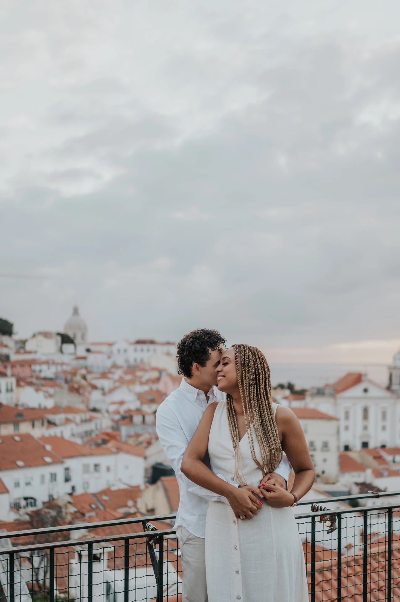 The man embracing his partner with the red rooftops and water behind them at the viewpoint during their couple photoshoot in Lisbon.