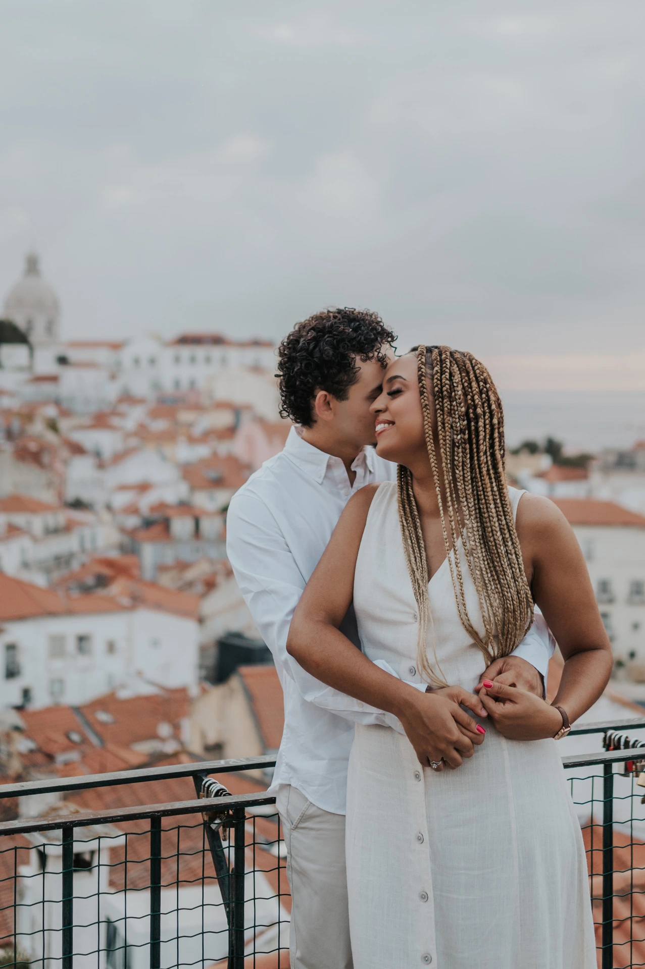 The man embracing his partner with the red rooftops and water behind them at the viewpoint during their couple photoshoot in Lisbon.