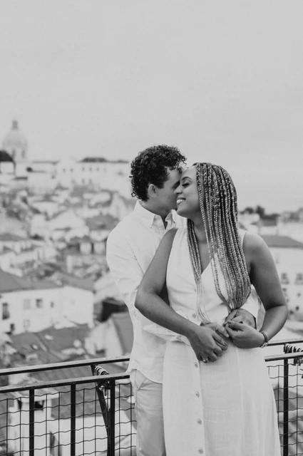 An embrace above the city in black and white | Lisbon couple photoshoot Black-and-white photograph: the man embracing his partner with the red rooftops and water behind them at the viewpoint during their couple photoshoot in Lisbon.