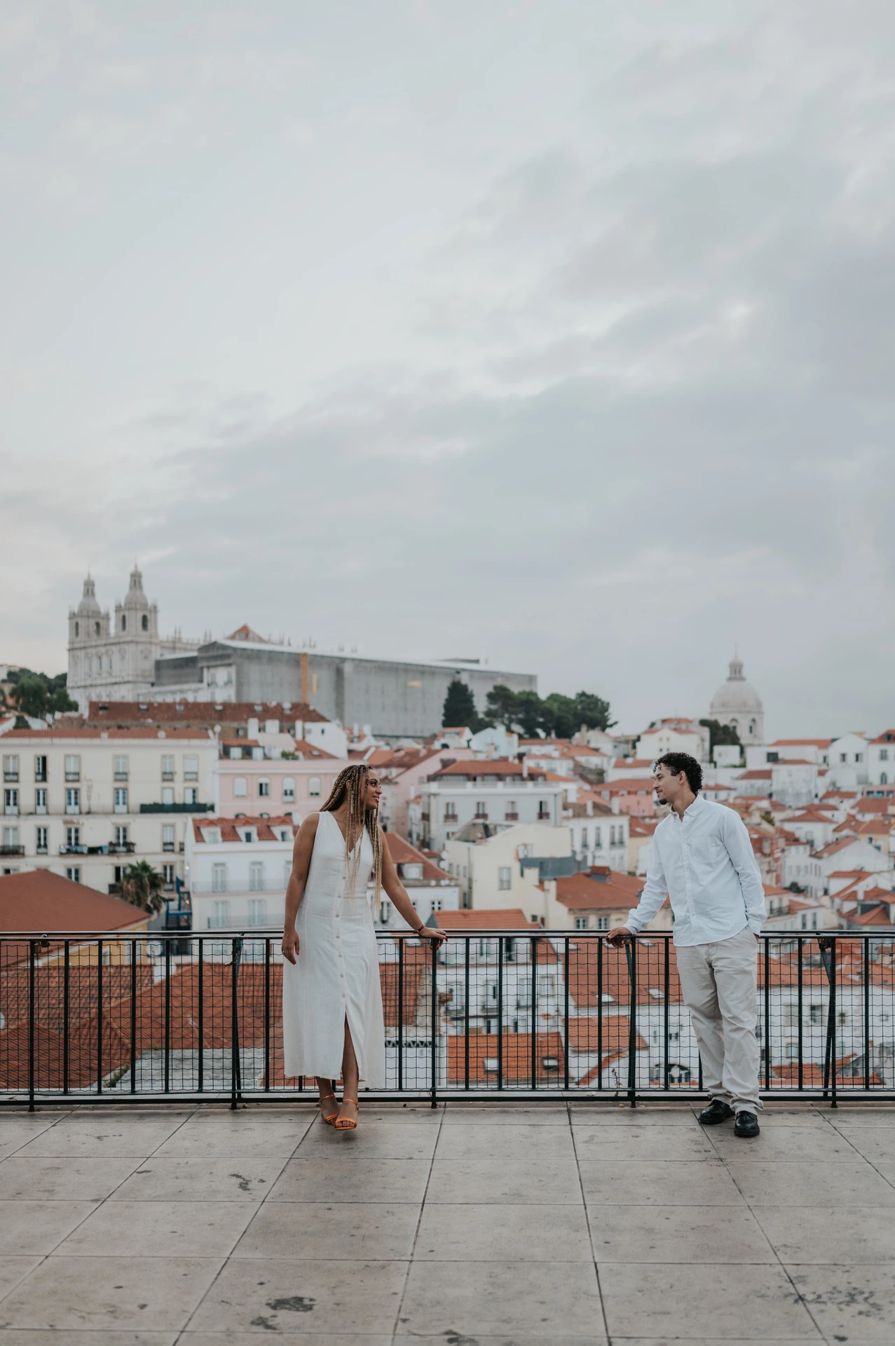 The couple holding hands beside the viewpoint railing with Lisbon’s rooftops behind them during their couple photoshoot in Lisbon.